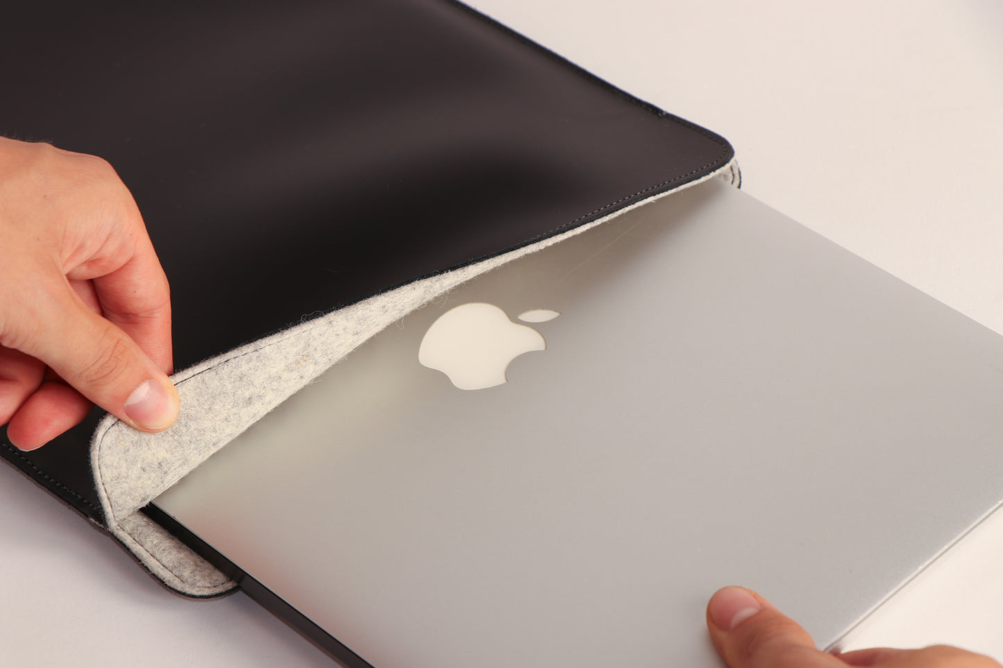 A close-up of a black leather laptop sleeve with a gray felt lining, shown being opened to reveal a silver Apple MacBook inside. A person's hands are visible holding the sleeve open.