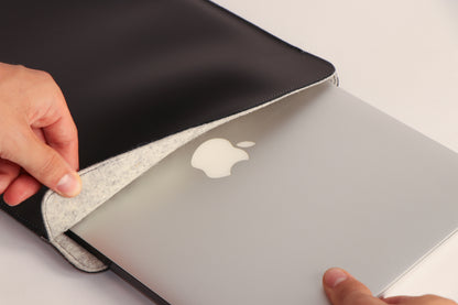 A close-up of a black leather laptop sleeve with a gray felt lining, shown being opened to reveal a silver Apple MacBook inside. A person's hands are visible holding the sleeve open.