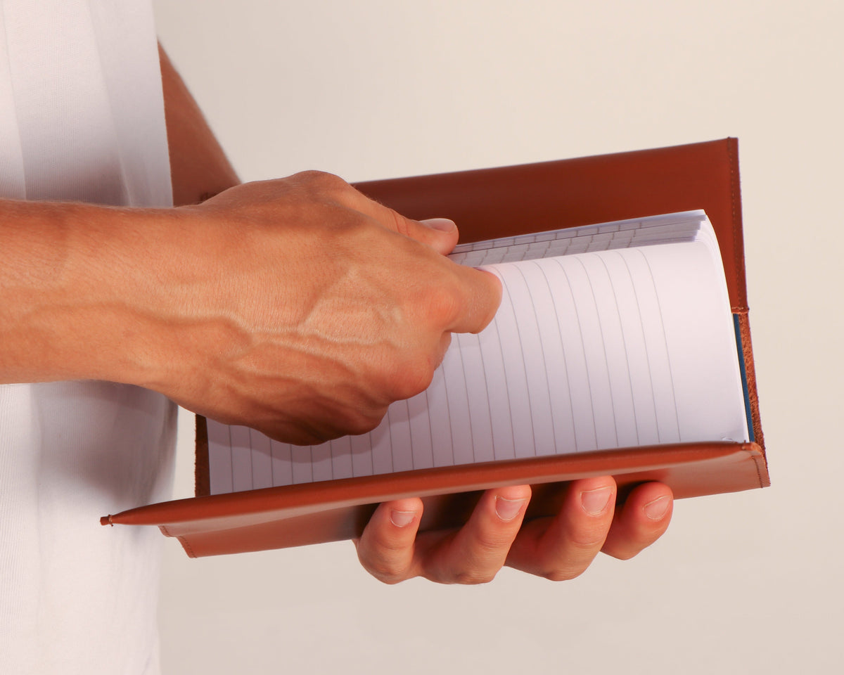 A close-up of a person's hands holding an open brown leather-bound notebook with lined white pages against a plain light background.
